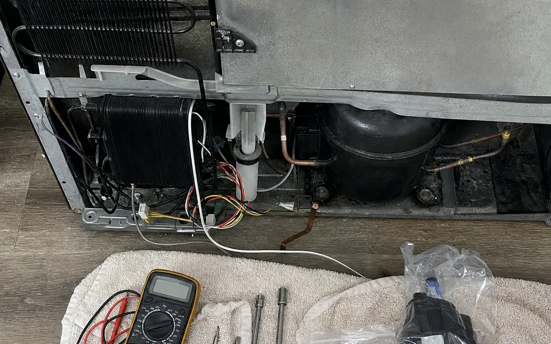 Machine compartment with the condenser coil, white drain tube and compressor exposed, a multimeter and tools on a towel in front.